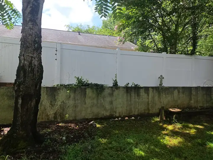 Concrete retaining wall covered in algae and mildew beneath a white vinyl fence, before pressure washing in Woodstock, GA.
