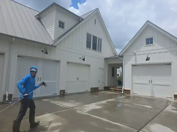 Technician pressure washing a concrete driveway in front of a white board-and-batten home with a three-car garage in Milton, GA.