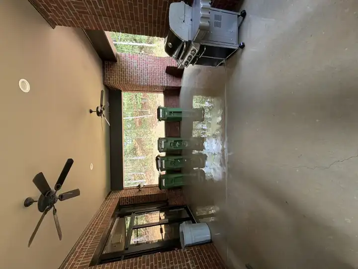 Wide covered patio during pressure washing in Alpharetta with standing water on the concrete floor, outdoor grill and trash bins visible along the back wall