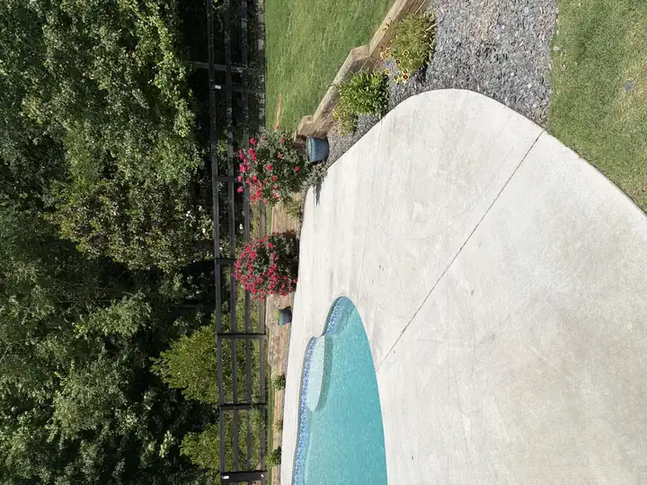 Freshly cleaned concrete pool surround in Canton, GA, with flower beds, stone retaining wall, and black wood fence in background